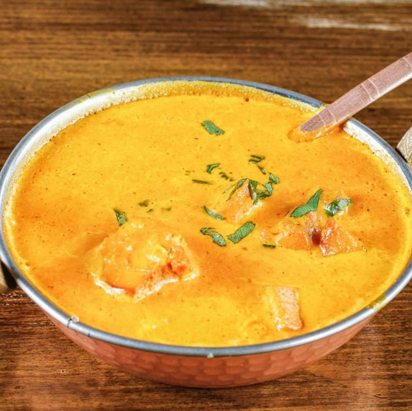 A copper bowl filled with creamy orange Indian curry, garnished with fresh green herbs, on a wooden table.
