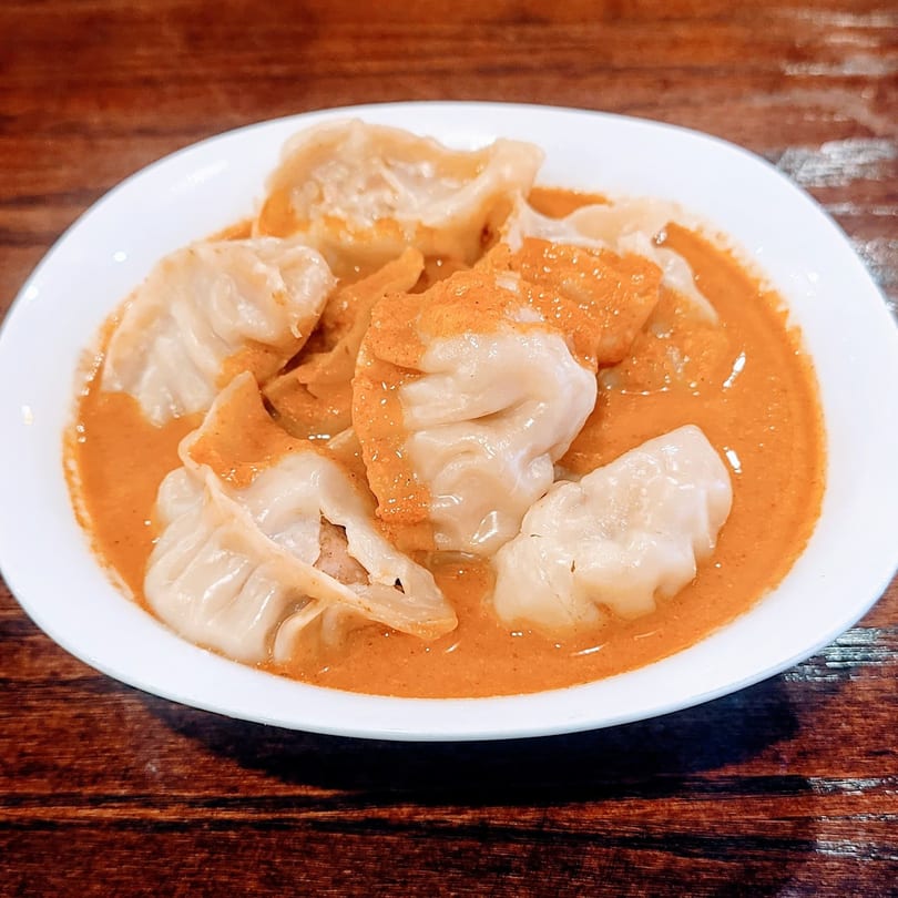 A white bowl of Nepali momos in a rich orange curry sauce, dusted with spices.
