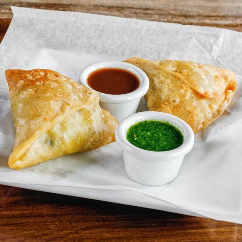 Two golden-brown samosas with small white bowls of green and red chutneys on a white paper-lined plate.