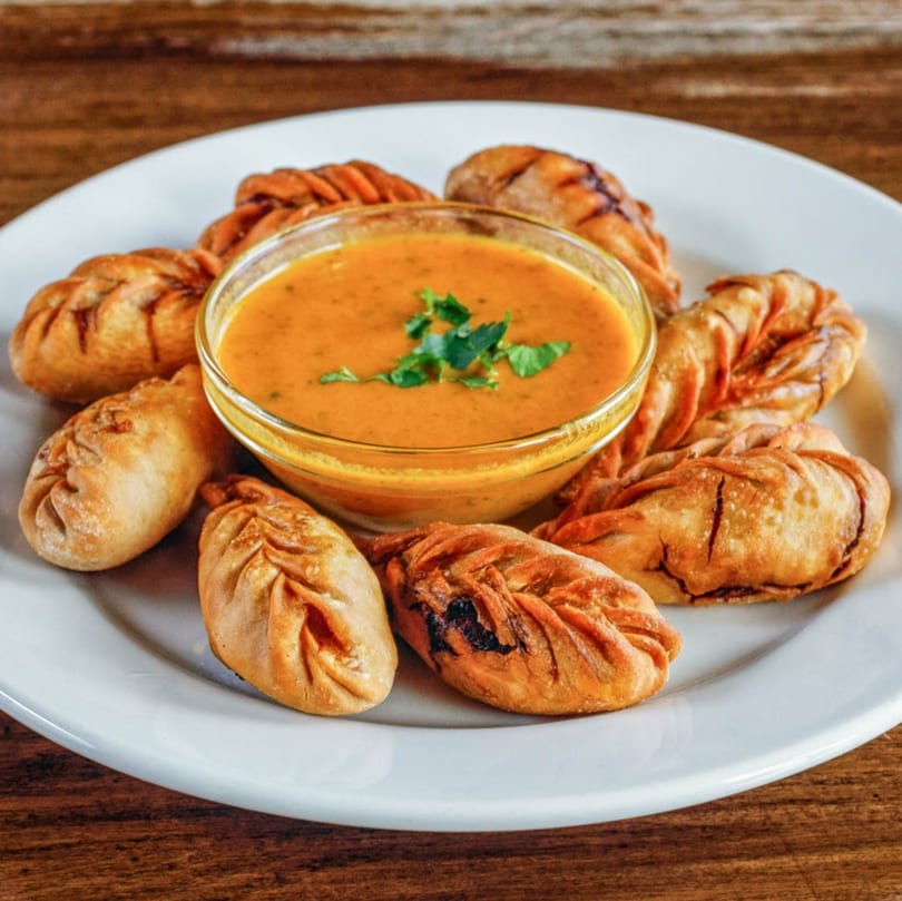 Fried momos, a Nepali dumpling, arranged around a bowl of orange dipping sauce on a white plate.